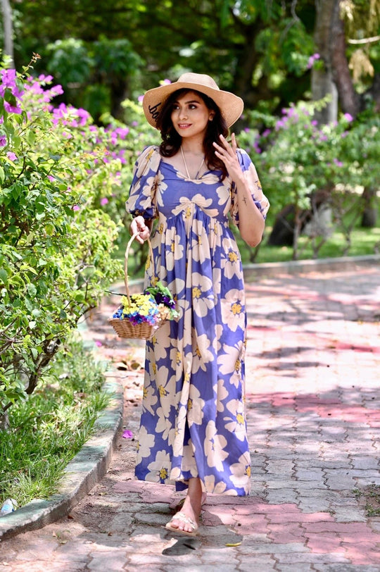 Woman in a floral dress and hat standing on a garden path with flowers and plants around.