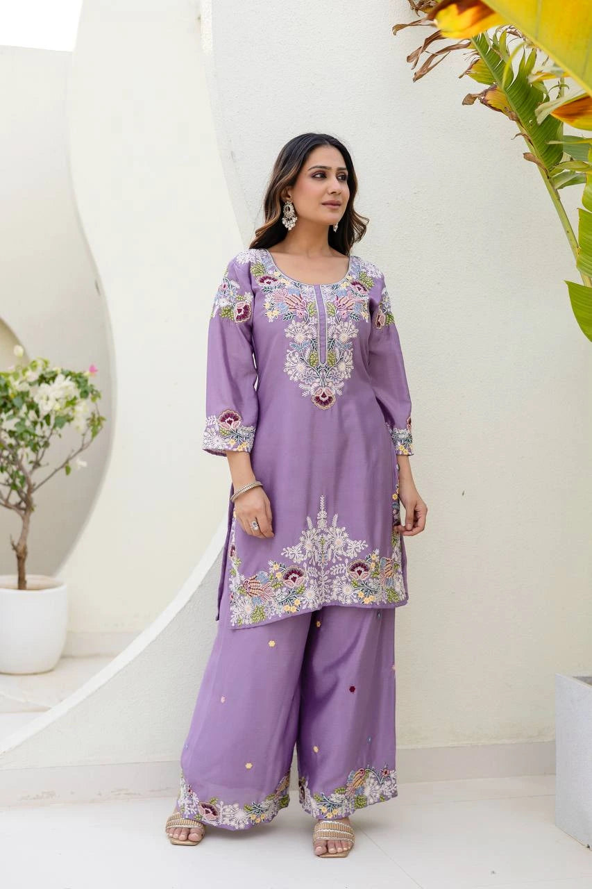 Woman wearing a purple embroidered outfit standing against a white wall with decorative elements.