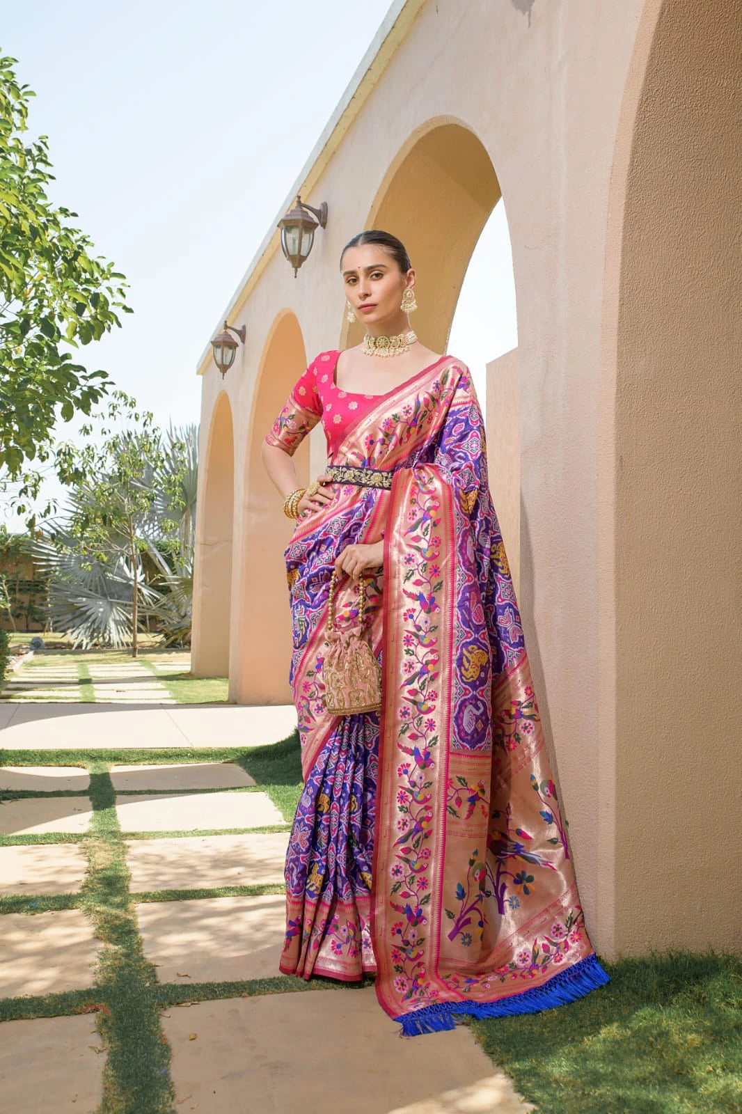 Woman in a colorful saree standing outdoors near a building with greenery.