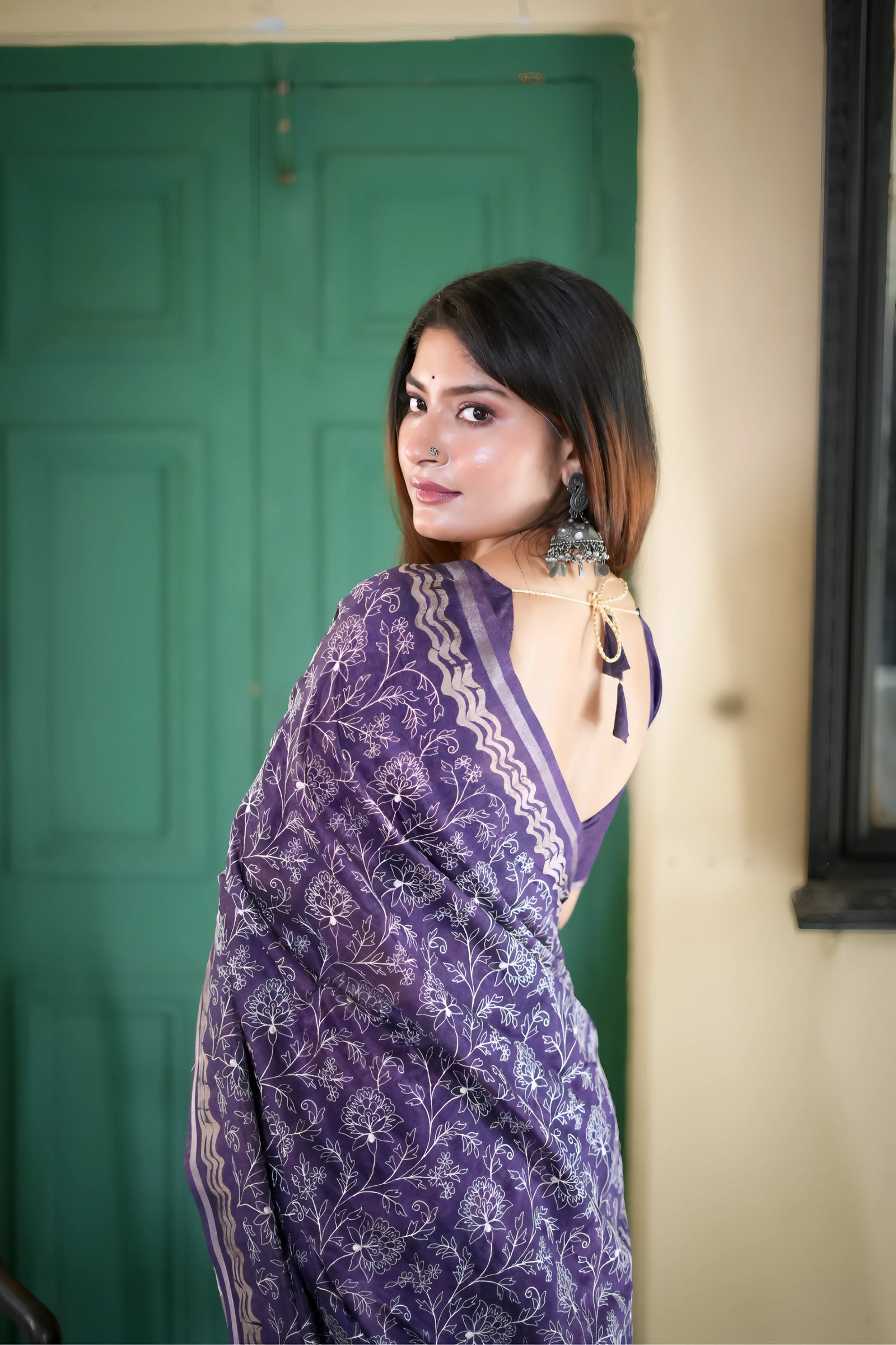 A woman models an elegant Deep Purple Tussar Silk Saree. The saree features dense all-over floral embroidery/weaving in a light metallic thread, framed by a contrasting metallic border. She wears a matching purple blouse and accessorizes with heavy oxidized silver jhumka earrings and bangles, posing indoors next to a teal door.