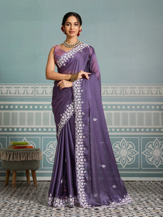 Woman wearing a purple saree with white embroidery against a decorative wall.