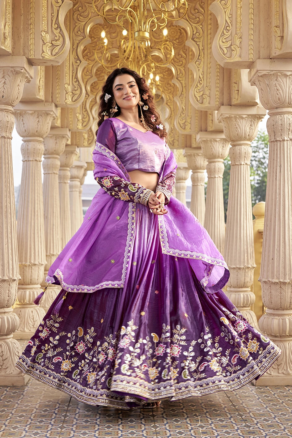 Woman in a purple traditional outfit with floral patterns standing in an ornate architectural setting.