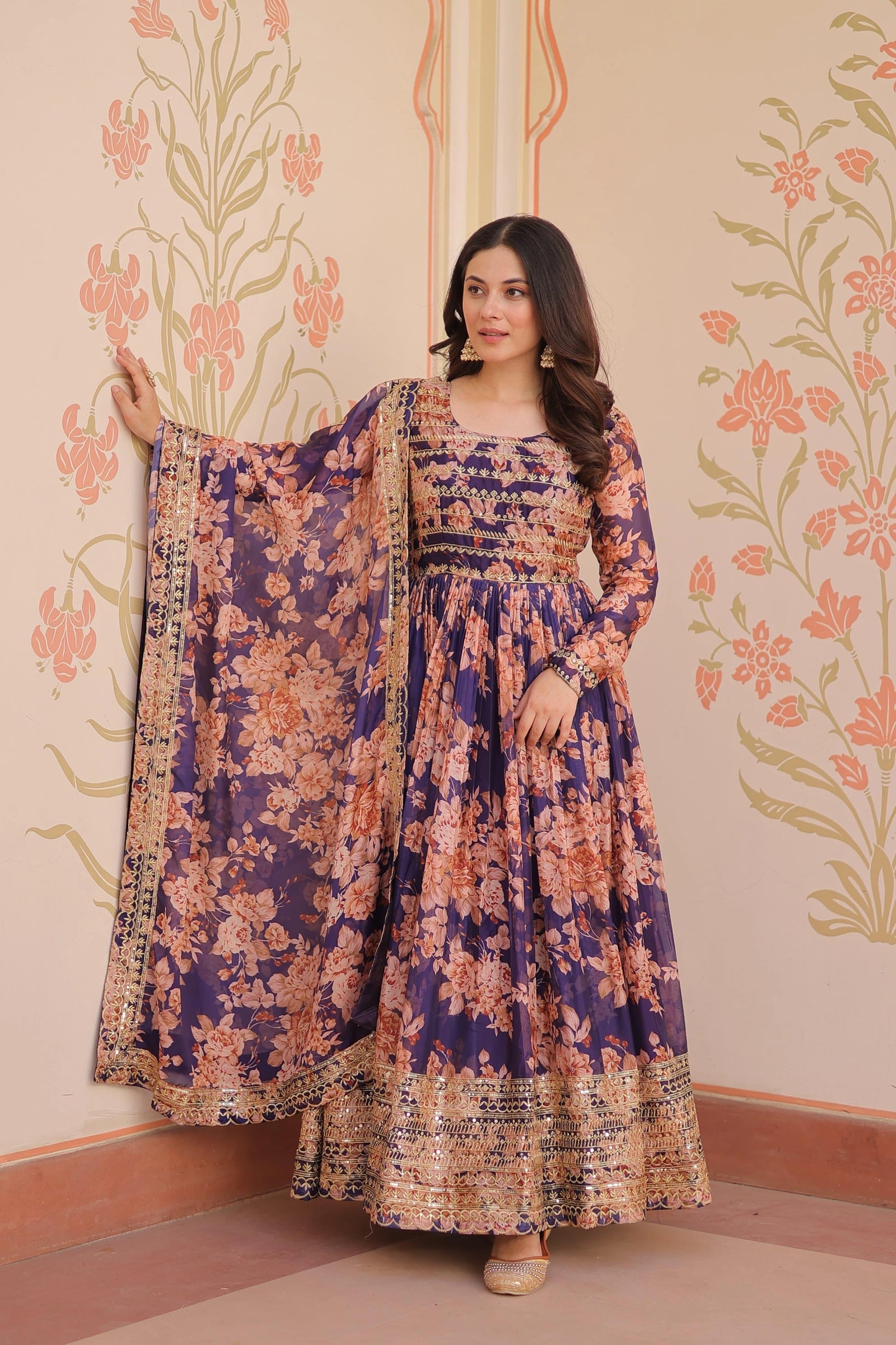 Woman wearing a floral dress with a matching dupatta against a decorative wall.