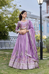 Woman in a purple embroidered traditional outfit standing outdoors with greenery and a building in the background.