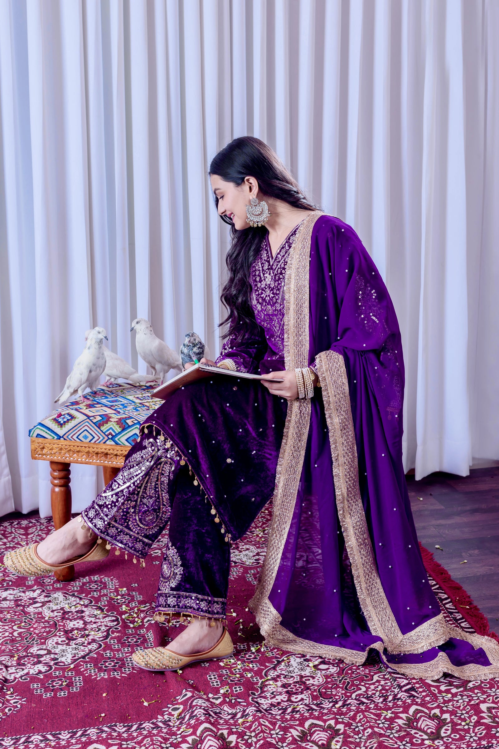 Woman in a purple traditional outfit sitting on a patterned rug with white curtains in the background.