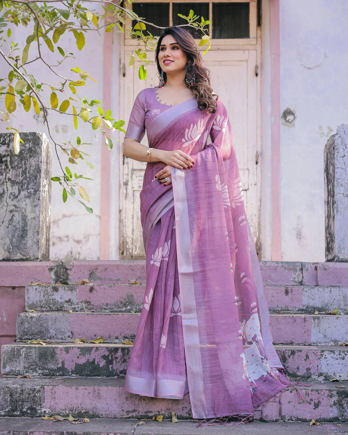 Woman in a purple saree standing outdoors with a white door and steps in the background.