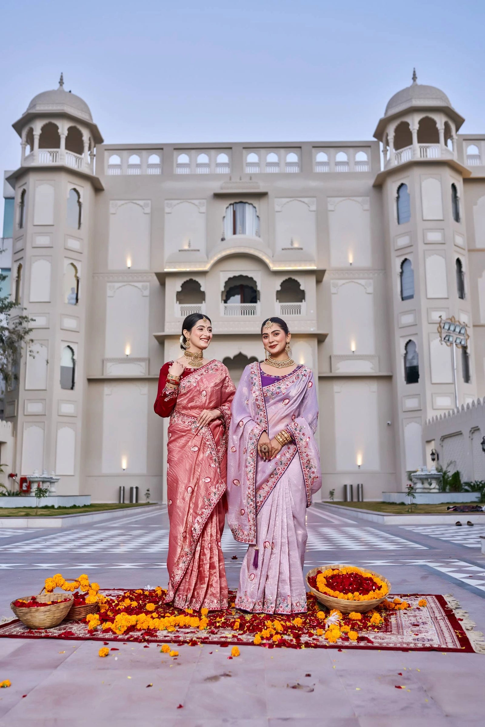 Two women in traditional sarees standing in front of a building with decorative elements on the ground.