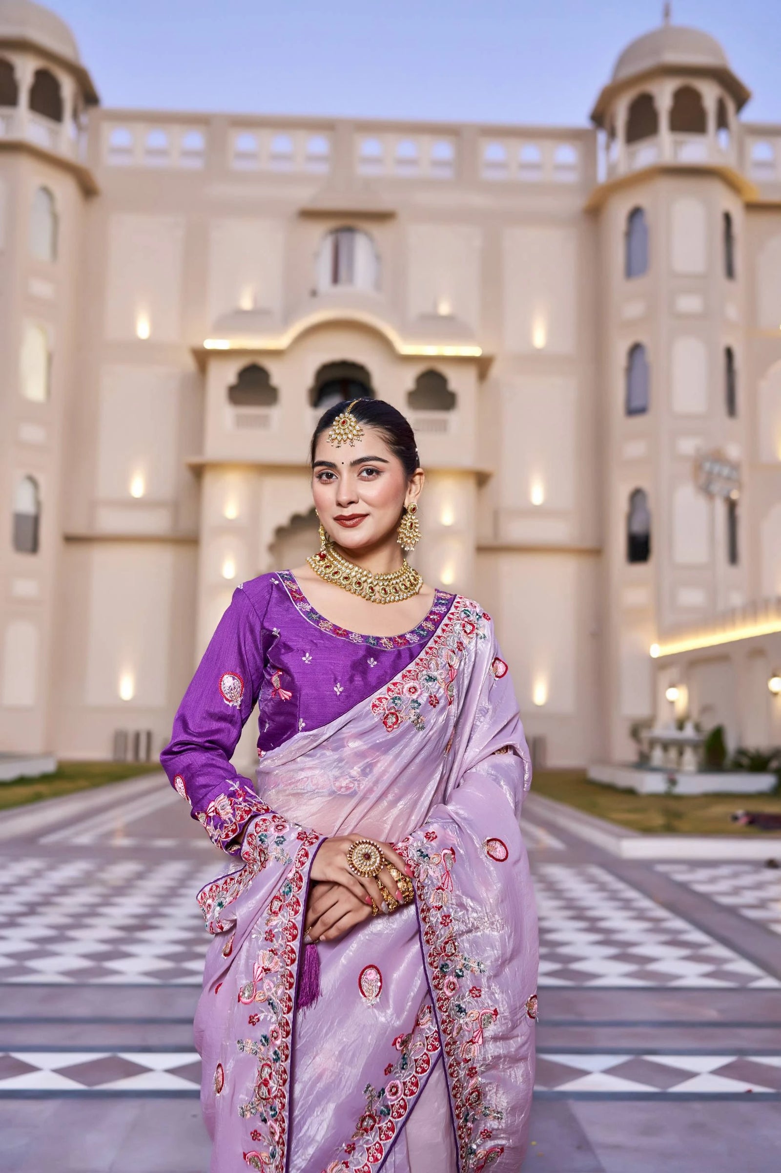 Woman in a traditional purple and pink saree standing in front of an architectural building.