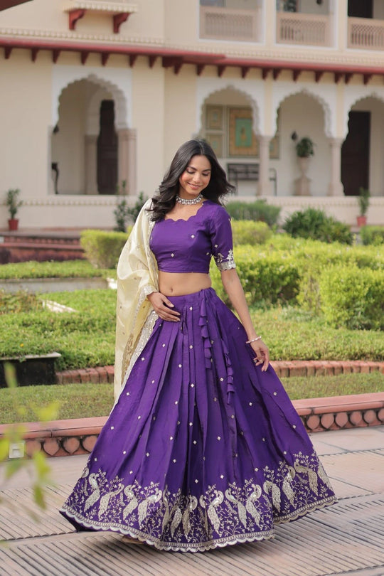 Woman in a purple traditional outfit standing in front of an architectural building with greenery.