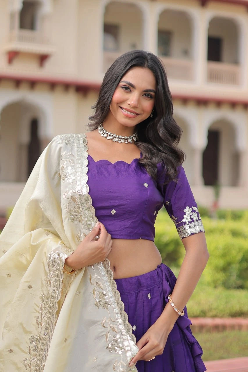 Woman in a purple blouse and white saree standing in front of a building with greenery.