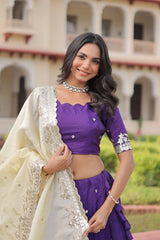 Woman in a purple blouse and white saree standing in front of a building with greenery.
