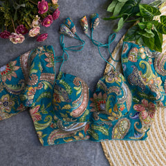 Teal padded blouse with intricate multicolor floral and paisley embroidery, sequin accents, and tie-back detail, displayed on a grey surface with artificial flowers and green leaves in the background.