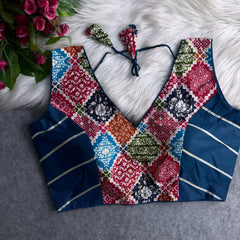 Traditional multicolor Ajrak-print blouse featuring striking geometric and floral motifs in navy blue, red, green, orange, and light blue. The blouse has a deep V-neckline, navy silk side panels with metallic silver stripes, and a matching tasseled tie-back, displayed flat on a white fur background with artificial pink flowers in the corner.