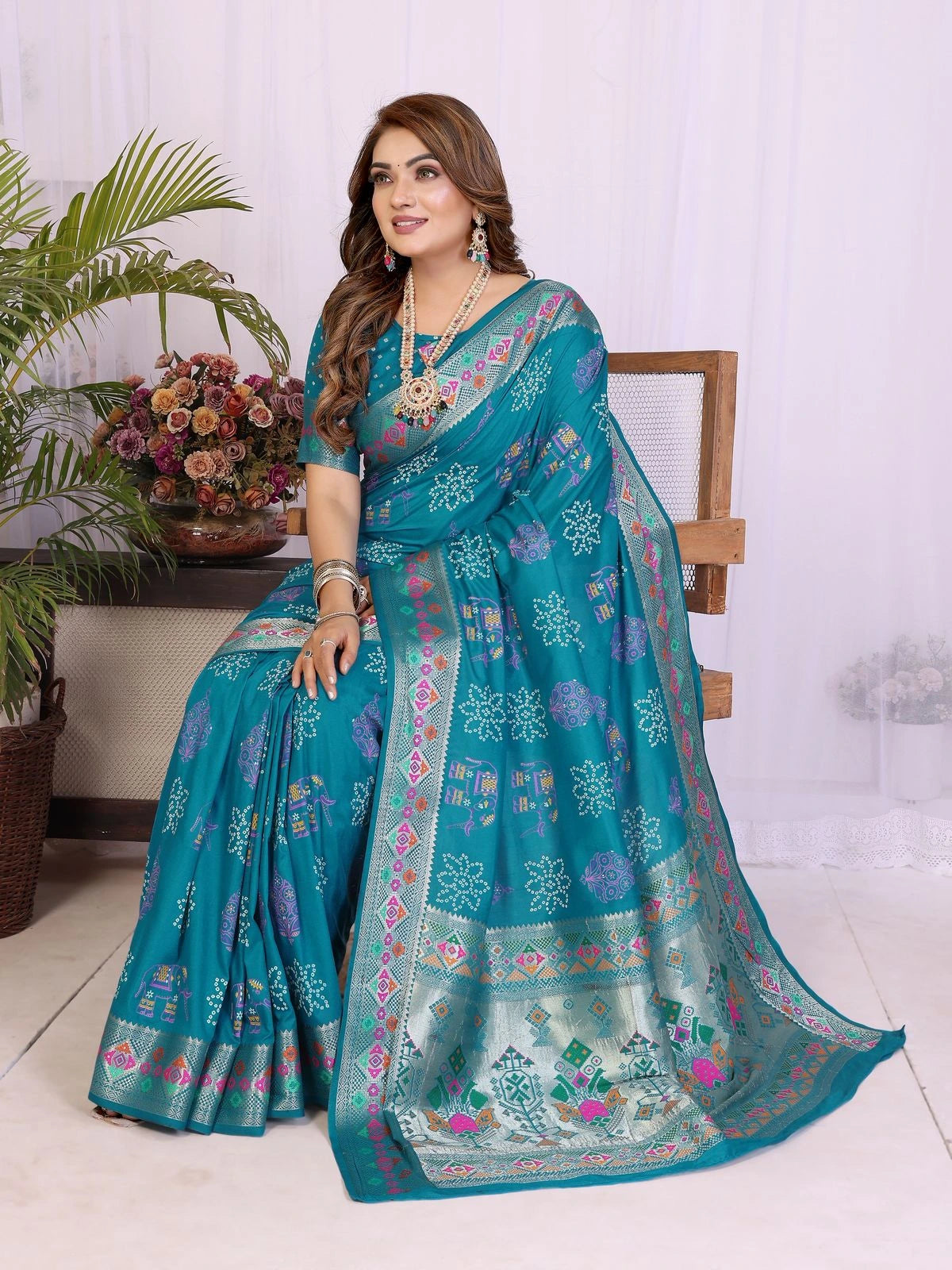 Woman wearing a teal saree with floral patterns in a room with plants and flowers.