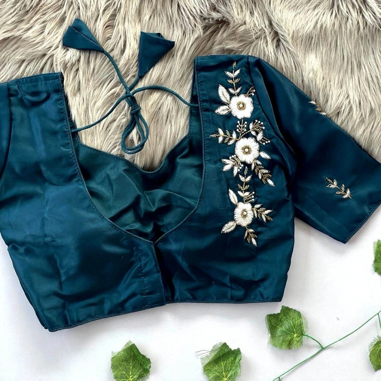 Teal blouse with floral embroidery on a white surface with green leaves.
