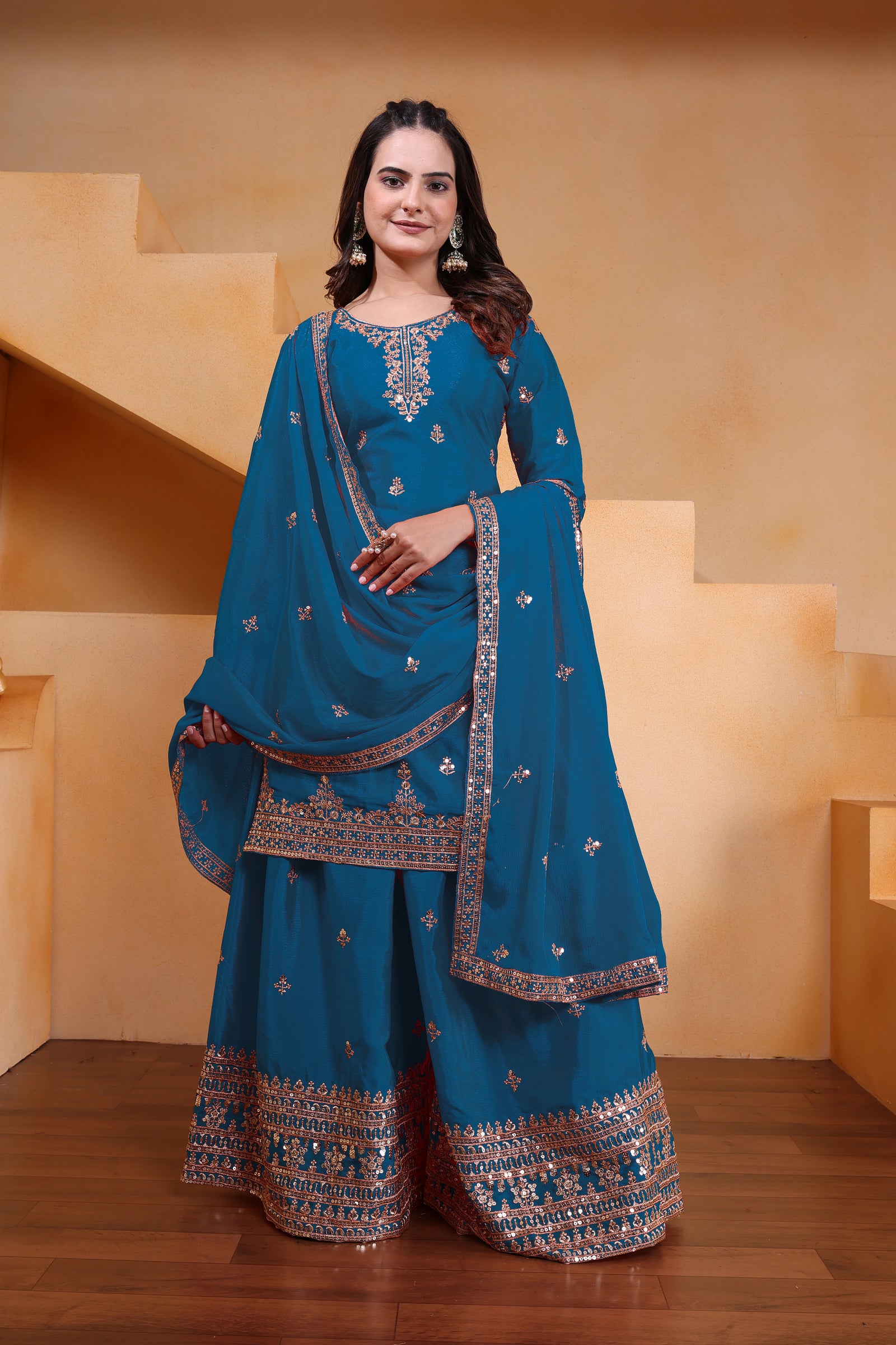 Woman in a blue traditional outfit with embroidery standing against a beige wall.