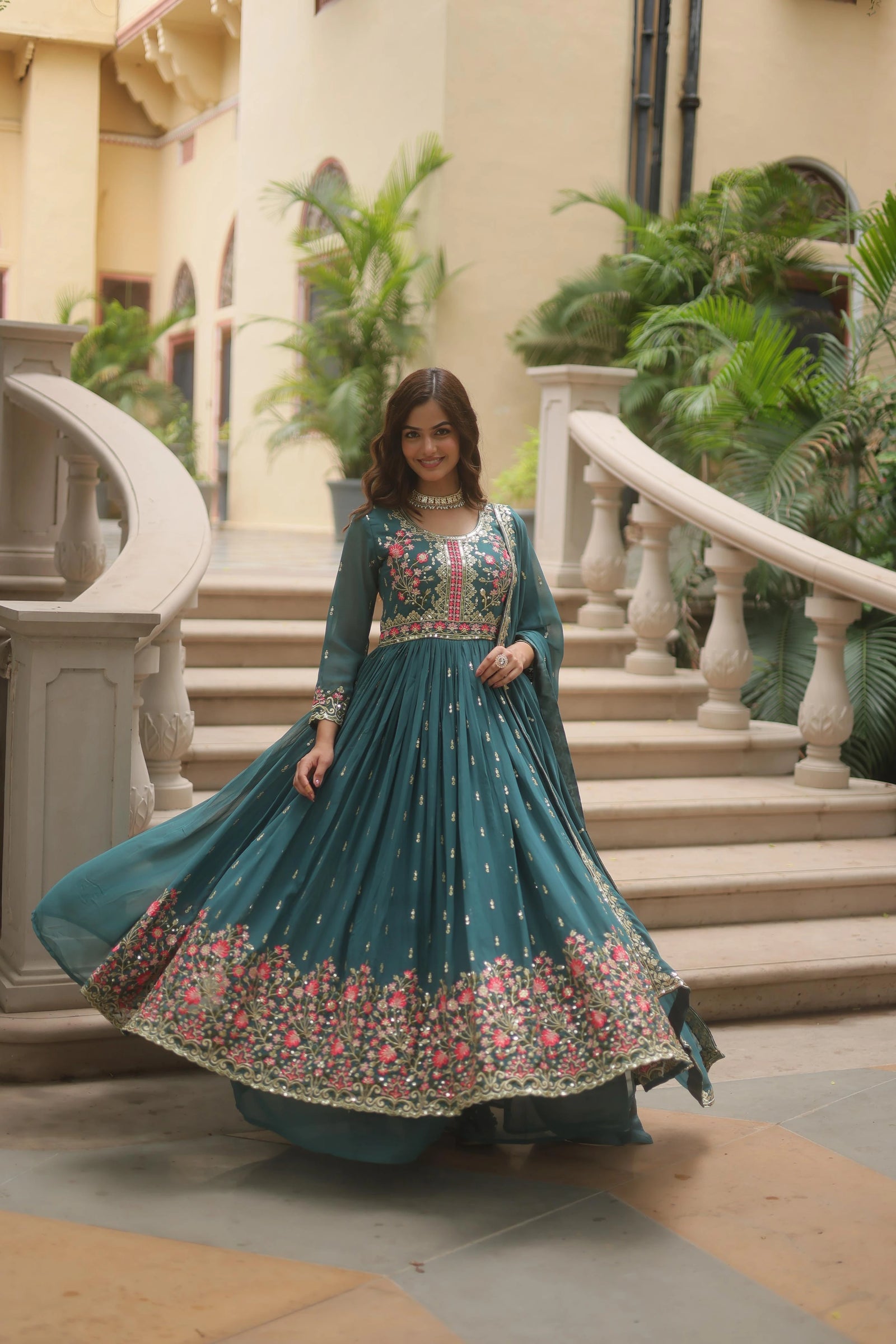 Woman in a teal embroidered dress standing in an elegant indoor setting with plants and stairs.