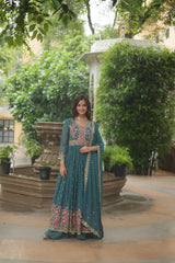 Woman in a teal traditional outfit with floral patterns standing outdoors near greenery.