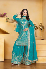 Woman in a teal embroidered traditional outfit standing in a decorative room.