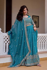 Woman in a teal traditional outfit with silver details standing in an elegant indoor setting.