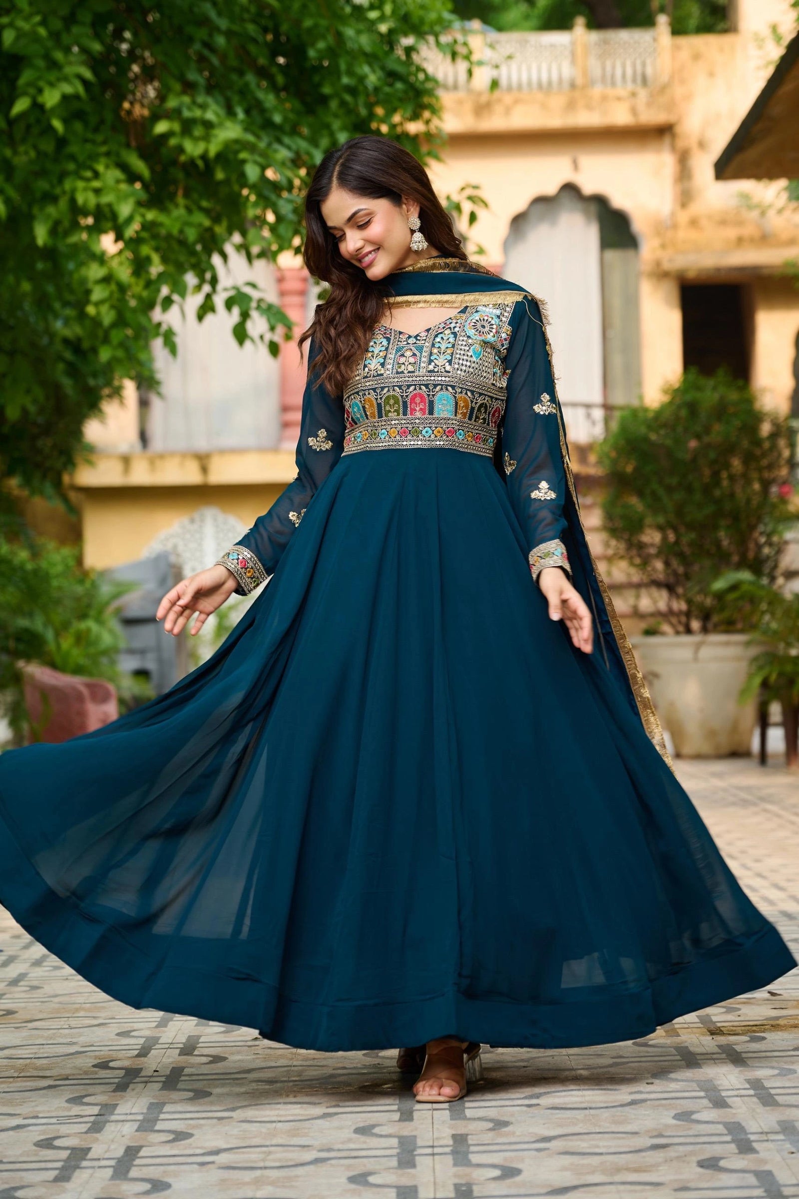 Woman in a teal embroidered traditional outfit standing outdoors with greenery and a building in the background.