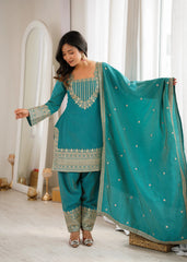 Woman in a teal traditional outfit with gold embroidery standing indoors.