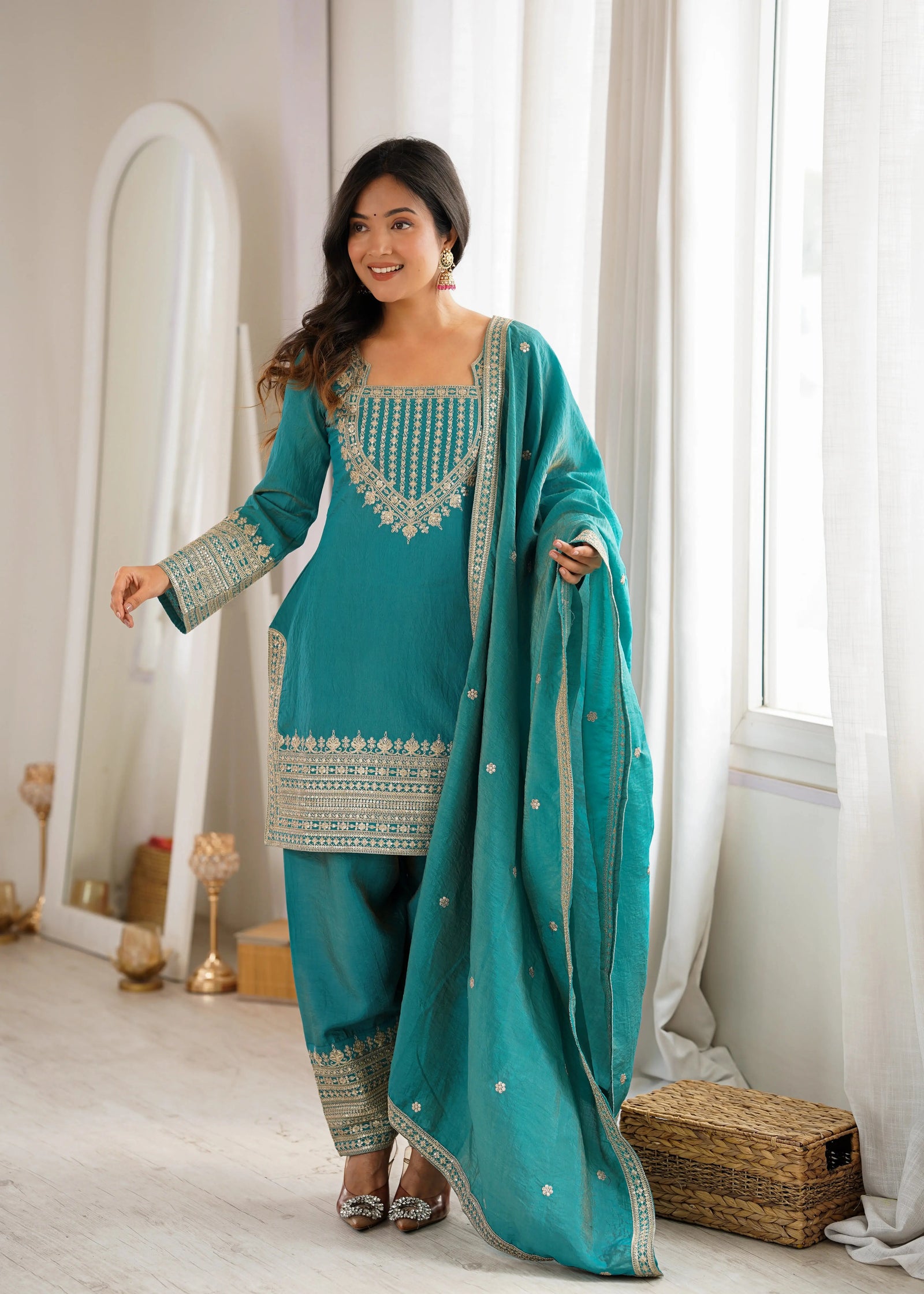 Woman in a teal traditional outfit standing in a bright room with large windows.