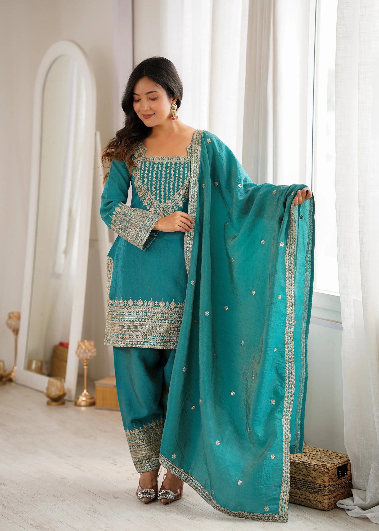 Woman in a teal traditional outfit with gold embroidery standing in a bright room.
