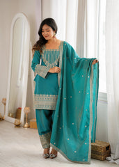 Woman in a teal traditional outfit with gold embroidery standing in a bright room.