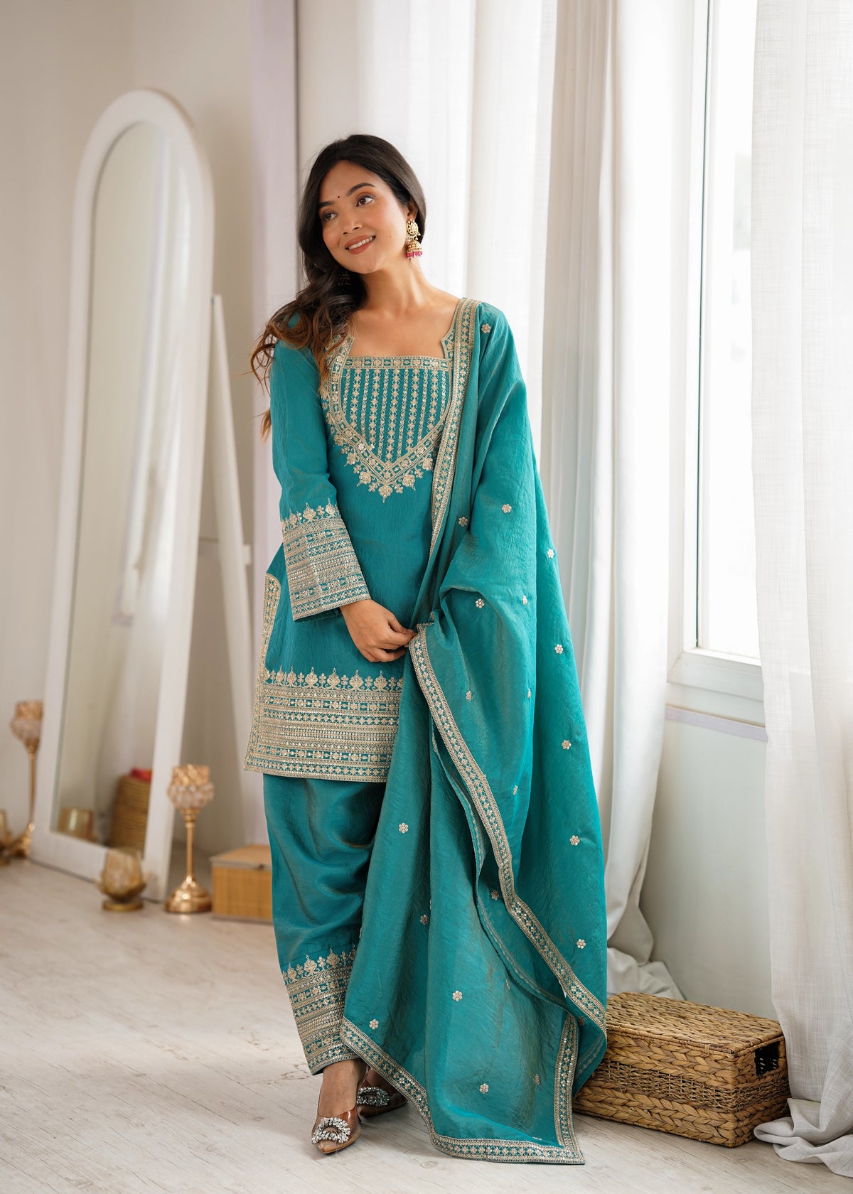 Woman in a teal traditional outfit standing in a room with white curtains and decor.