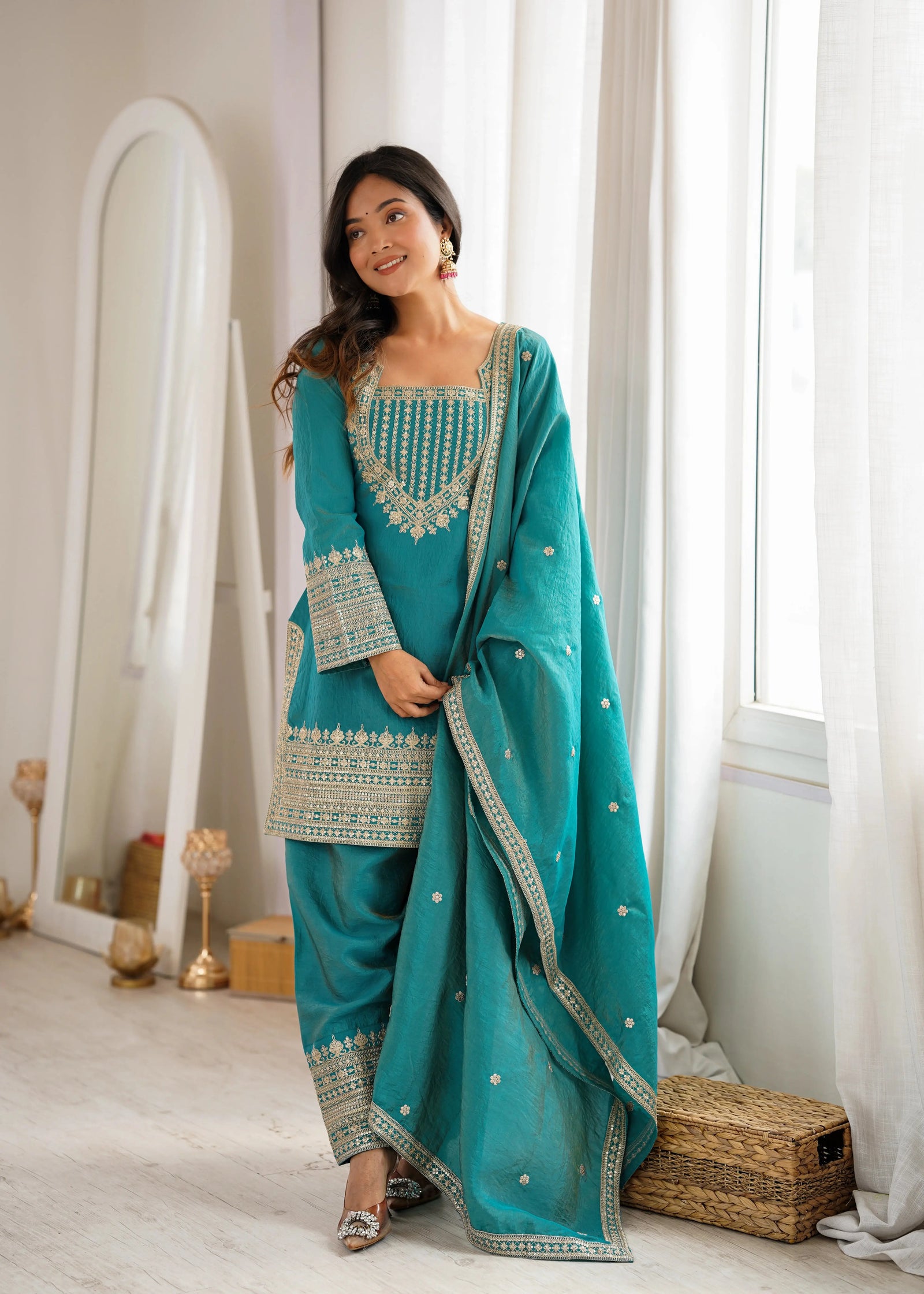 Woman in a teal traditional outfit standing in a room with white curtains and decor.