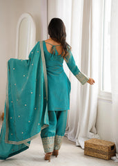 Woman in a teal traditional outfit standing in a room with white curtains.