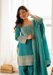 Woman wearing a teal traditional outfit with white embroidery indoors.