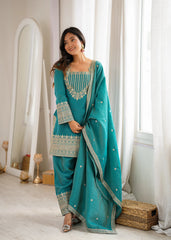 Woman in a teal traditional outfit standing in a room with white curtains and decor.