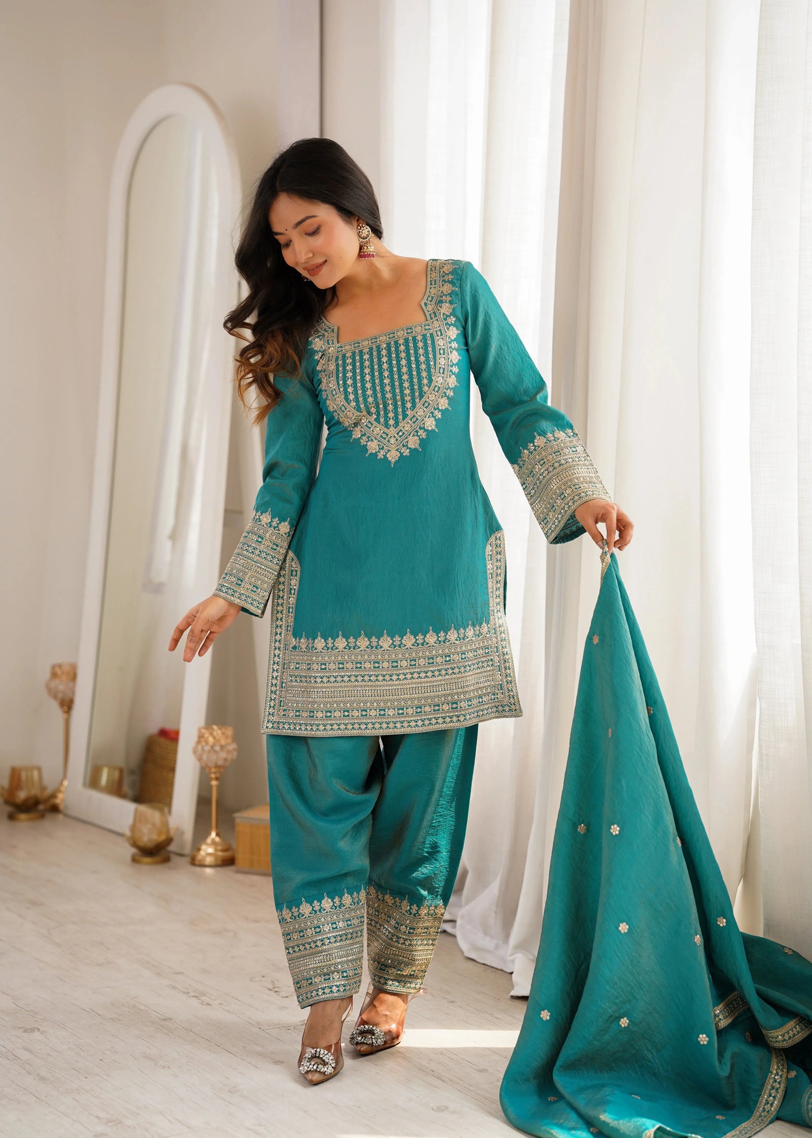 Woman in a teal embroidered traditional outfit holding a matching dupatta in a well-lit room.