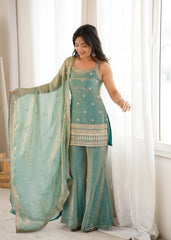 Woman in a traditional teal outfit with a matching dupatta standing in a bright room.