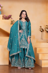 Woman in a teal traditional outfit with silver embroidery standing indoors.
