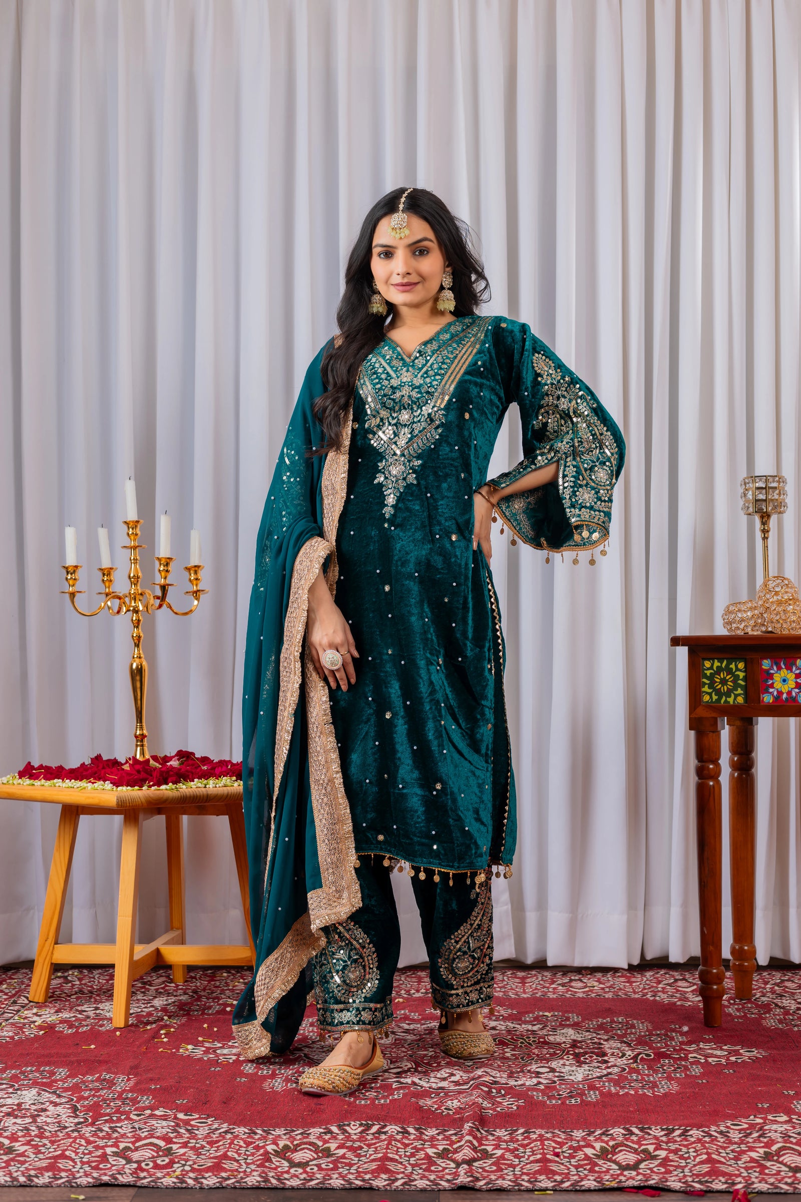 Woman in a teal traditional outfit standing in a decorated room with a white curtain and red carpet.