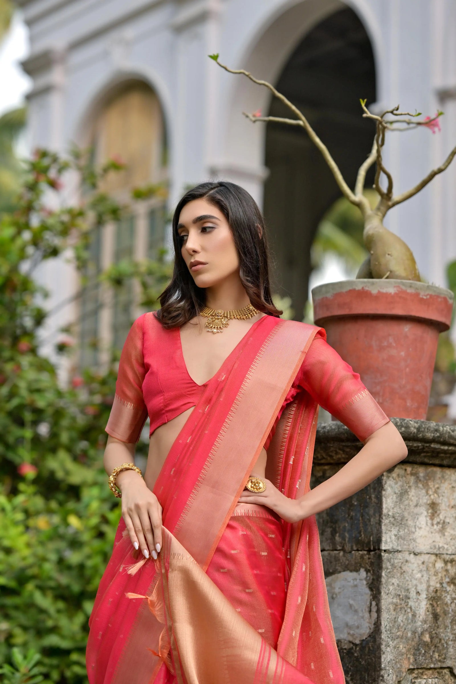 A woman models a stunning Coral Red Soft Silk Saree. The sheer saree features subtle metallic motifs and a wide, contrasting metallic gold/bronze woven border and pallu. She wears a matching red short-sleeve blouse and delicate gold jewelry, posing gracefully on an ornate stone staircase.