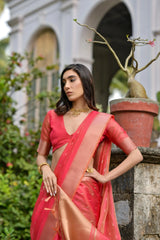 A woman models a stunning Coral Red Soft Silk Saree. The sheer saree features subtle metallic motifs and a wide, contrasting metallic gold/bronze woven border and pallu. She wears a matching red short-sleeve blouse and delicate gold jewelry, posing gracefully on an ornate stone staircase.