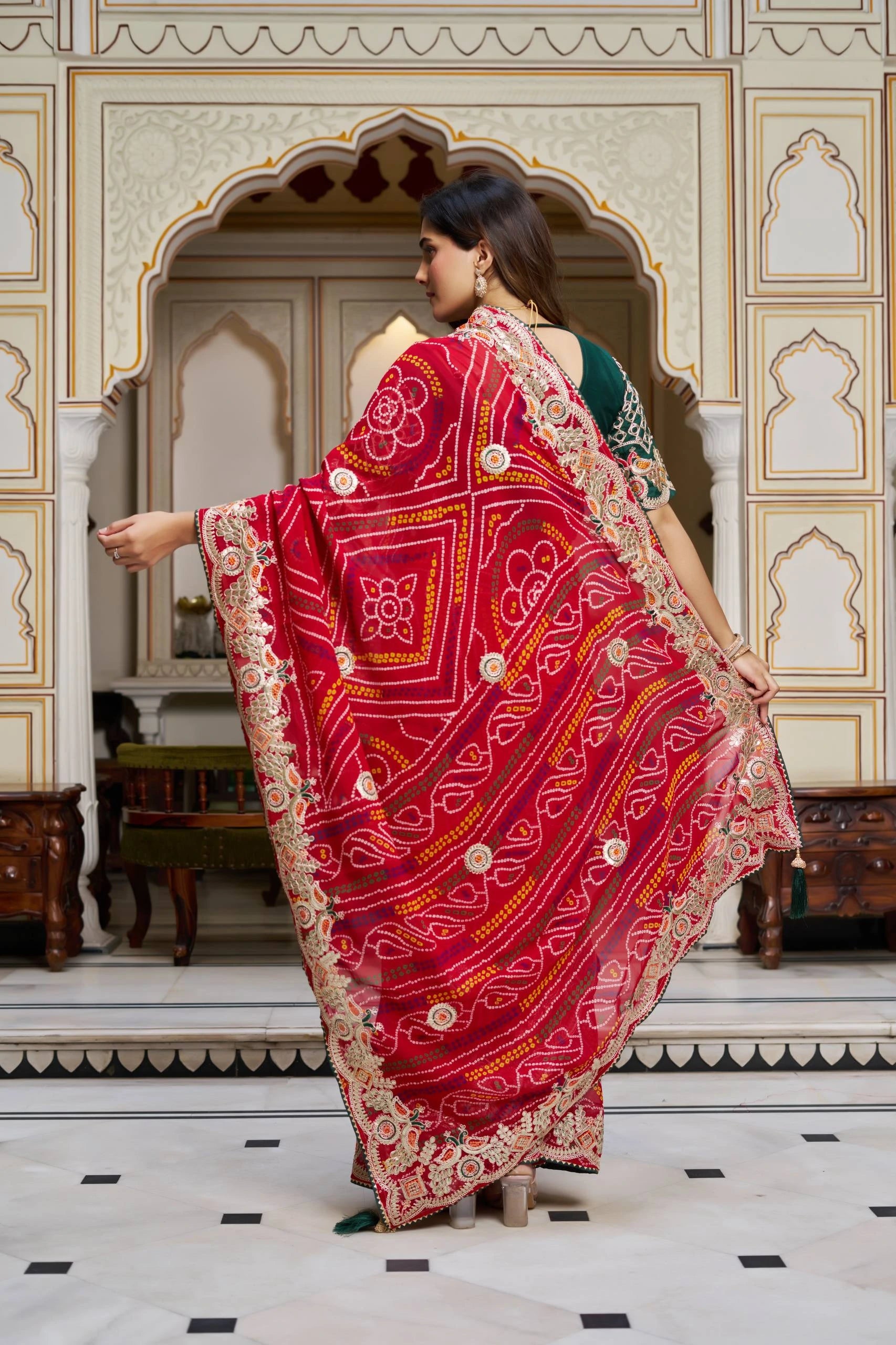 Woman in a red saree with gold patterns in an ornate room.