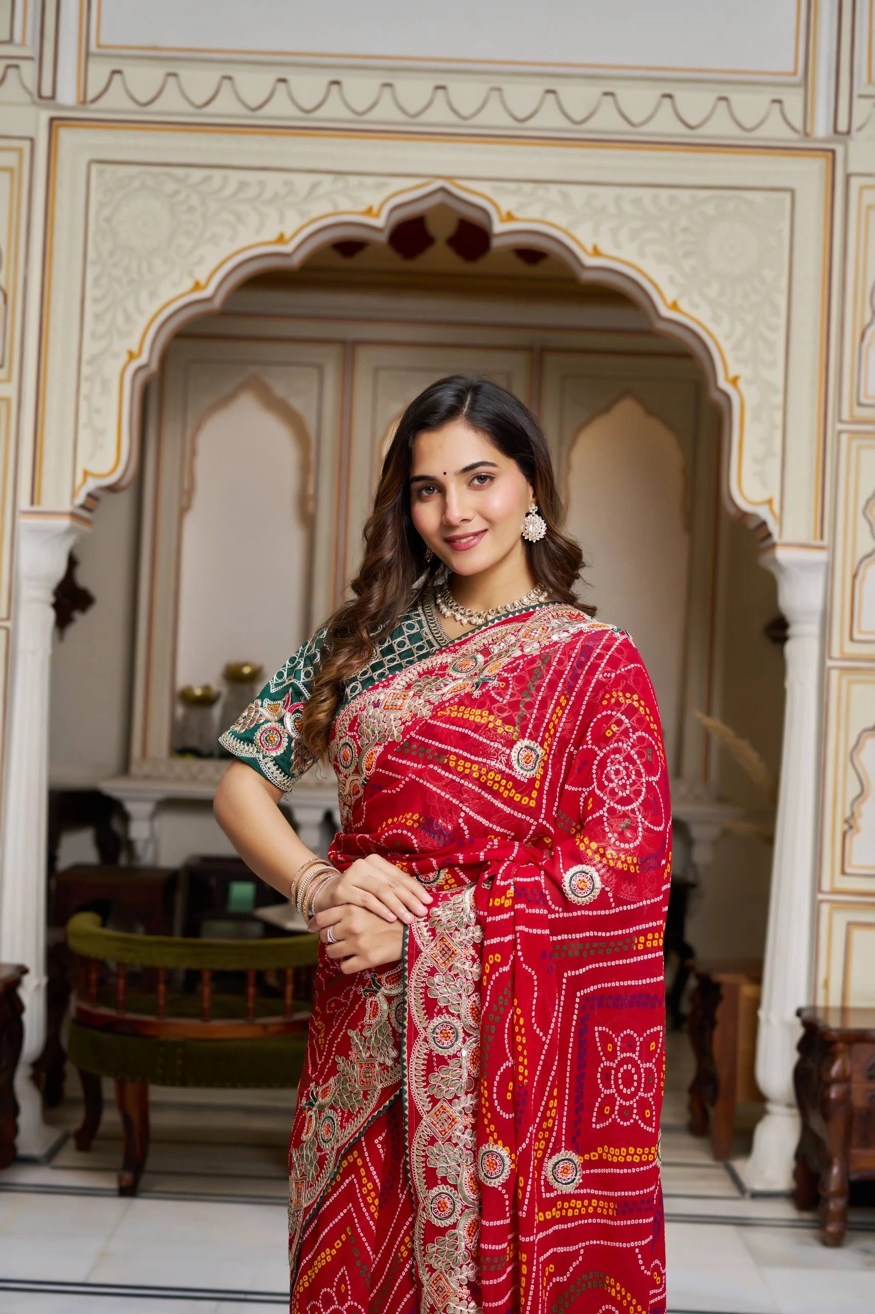 Woman in a red saree with intricate patterns standing in an ornate room.