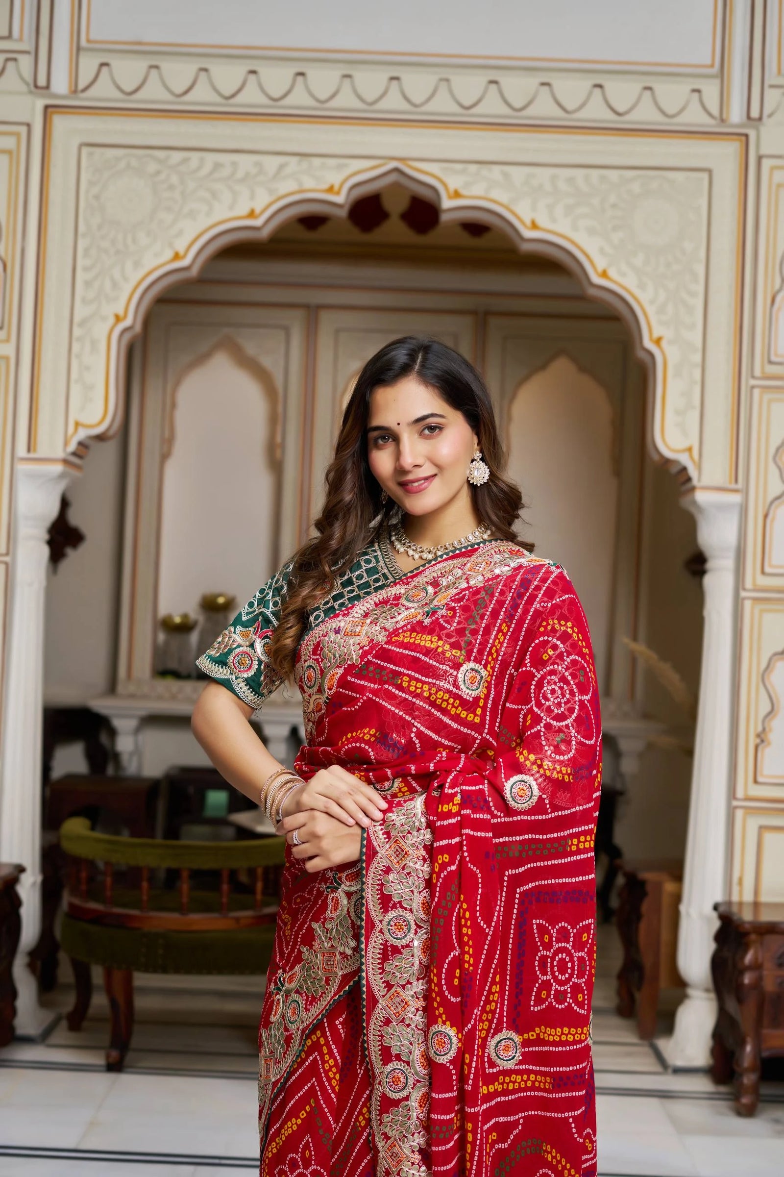 Woman in a red saree with intricate patterns standing in an ornate room.