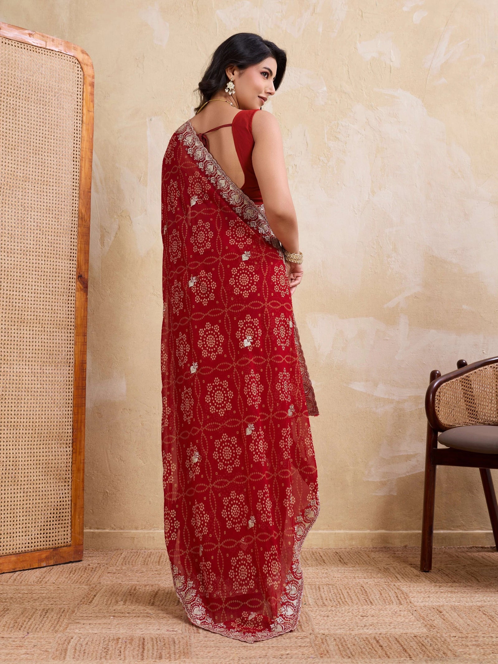 Woman wearing a red saree with white patterns in a room with a textured wall and wooden chair.