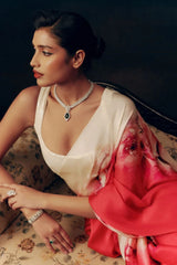 Woman in a white and red saree with jewelry, sitting on a patterned chair.