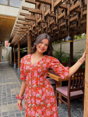 Woman in a red floral dress standing outdoors under a wooden architectural structure.