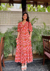 Woman in a red floral dress standing in front of green plants and a textured wall.