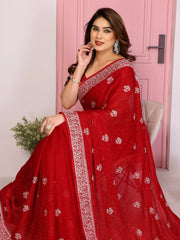 A medium shot of a woman sitting gracefully on a chair, draped in a red saree with detailed silver embroidery. She is smiling softly while looking toward the side, highlighting the delicate texture and shimmer of the fabric.