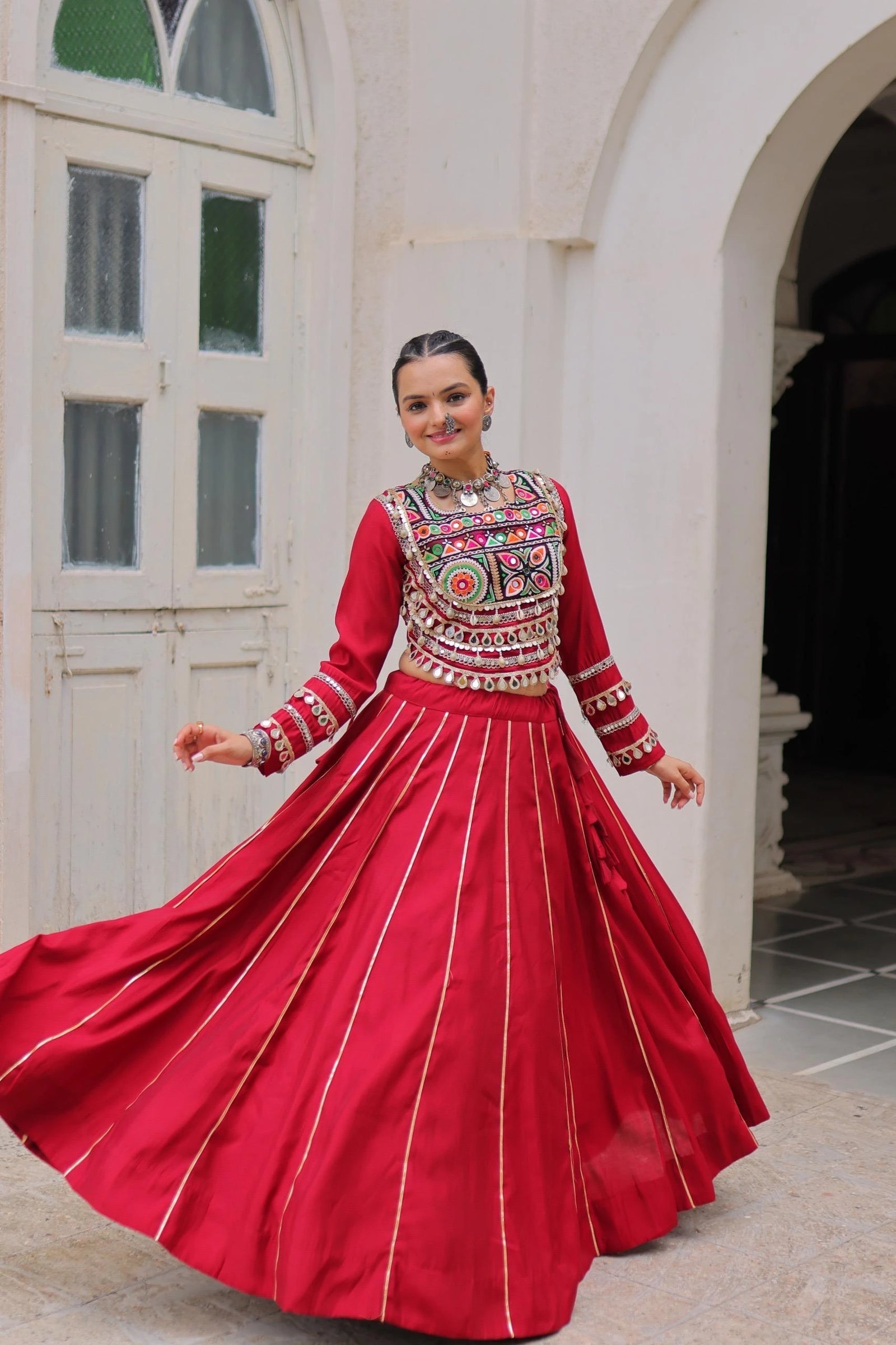 Woman in a traditional red outfit with intricate patterns standing in front of a white archway.