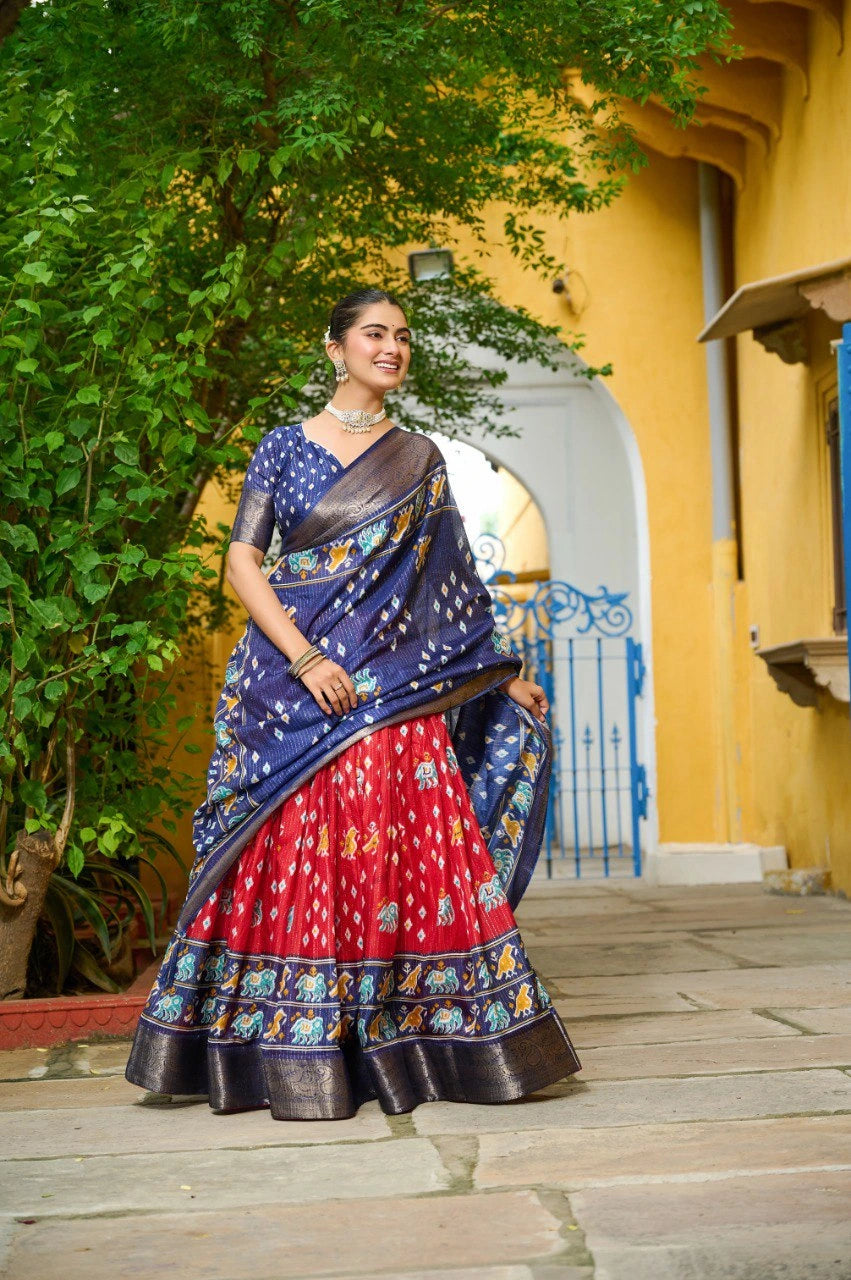 Woman in a colorful saree standing outdoors with a yellow building and greenery in the background